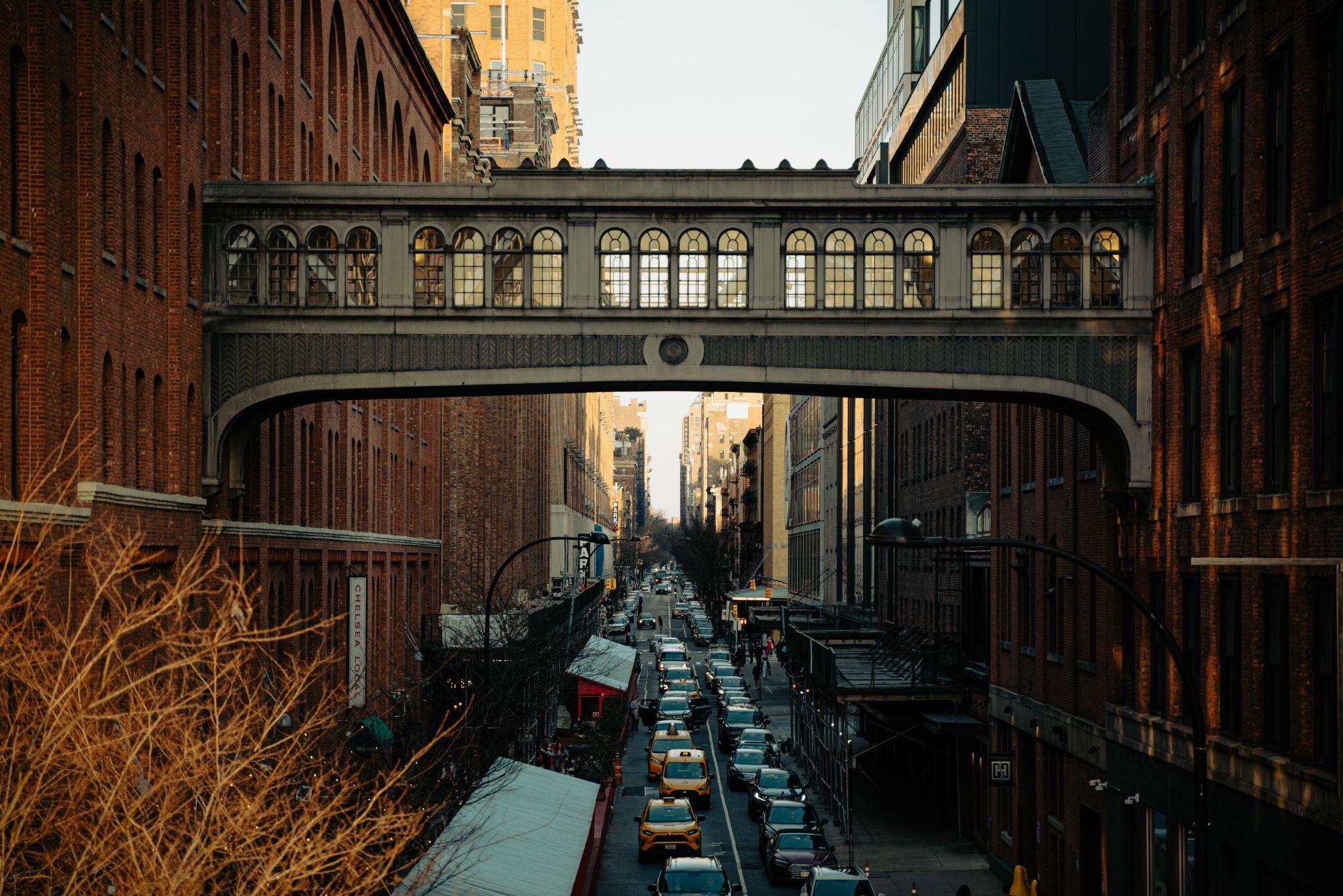 Looking down a Chelsea street from above: an arched stone skybridge spans two brick buildings over traffic and yellow cabs in golden-hour light.