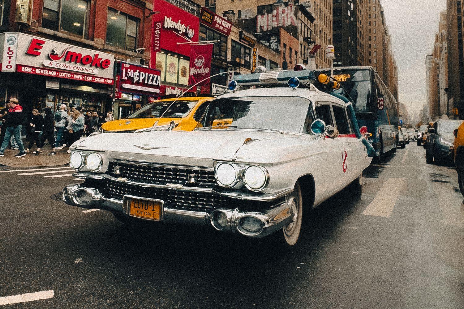 The white Ecto-1 Cadillac from Ghostbusters driving down a wet Manhattan avenue beside a yellow cab, storefronts and a crowd on the sidewalk behind.