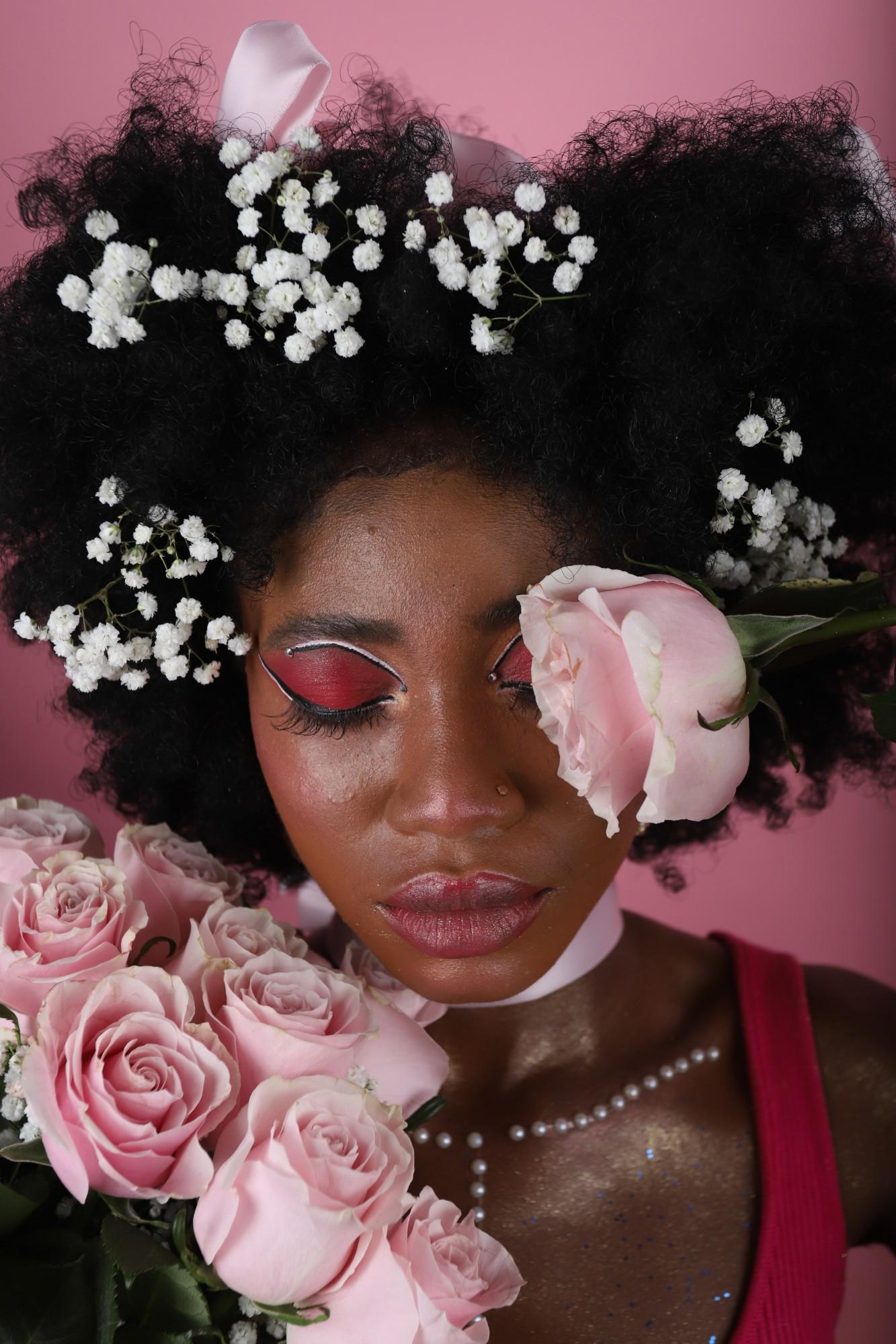 Portrait on a pink backdrop, eyes closed under red graphic eyeshadow, baby's breath in a full natural afro, one pink rose over an eye and a bouquet of pink roses below.